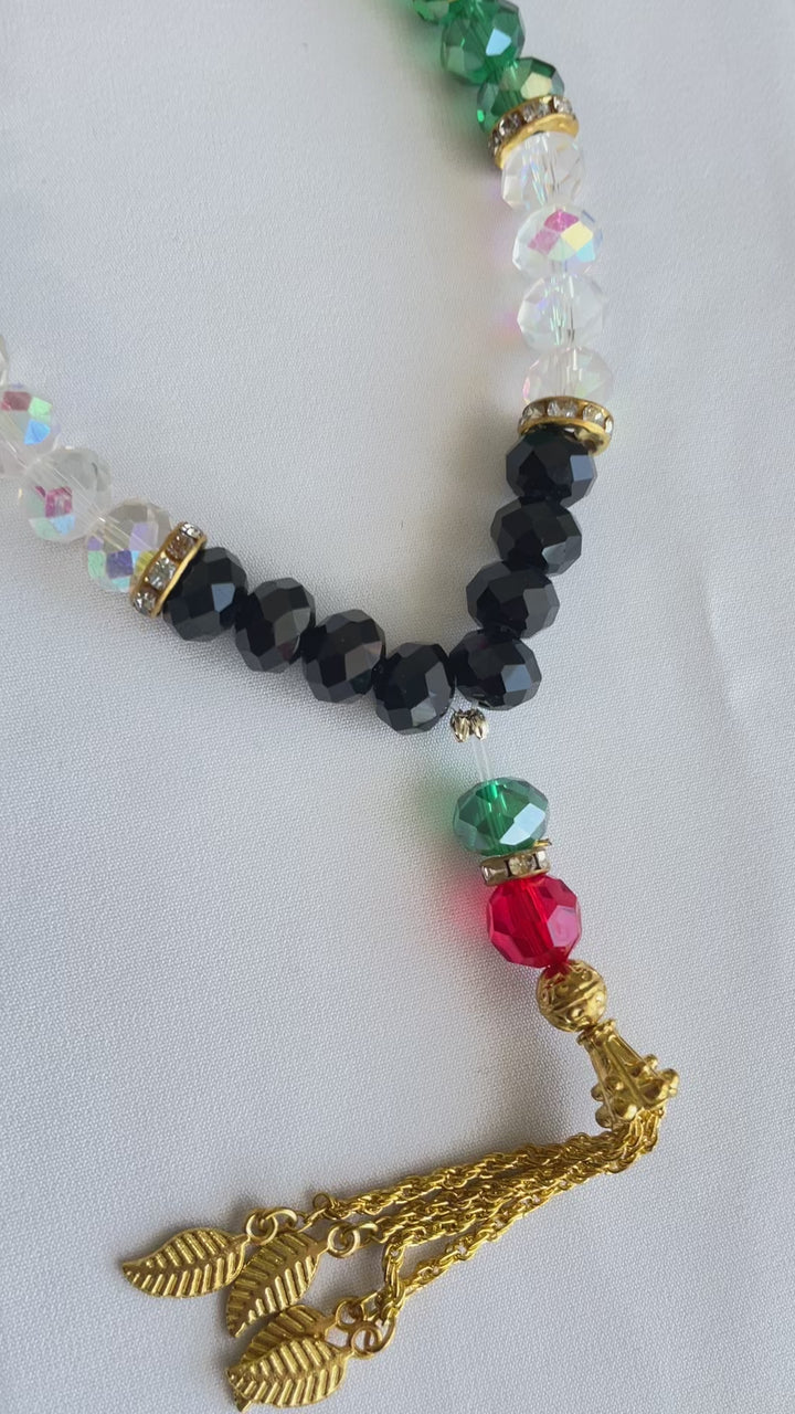 An elegant rosary featuring a design inspired by the colors of the Palestinian flag, where lustrous crystal beads in red, green, white, and black alternate, giving it a distinctive national character. Gold metallic spacers are interspersed between the beads, enhancing their sparkle and allure. The rosary is crowned with dangling golden metallic tassels, consisting of delicate chains ending with metallic leaf-shaped ornaments, adding a touch of luxury and sophistication to the rosary as a whole.